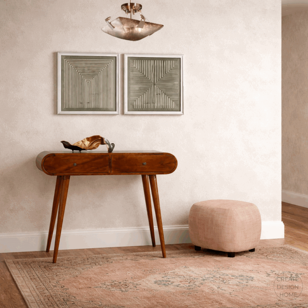 Transitional entryway featuring a curved wood console table, geometric wall art, a soft blush ottoman, and a muted patterned rug.